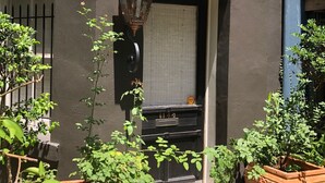 A building facade with a black door, a window with a closed shutter, and a metal gate. There are potted plants in front of the building.