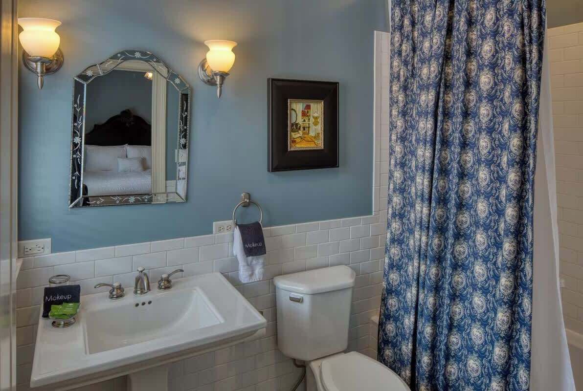 Un bagno con un lavandino bianco, un water, uno specchio con cornice decorativa, applique a parete, un quadro incorniciato e una tenda da doccia fantasia.