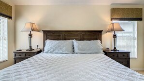 Una cama bien hecha con una colcha blanca acolchada, flanqueada por dos mesitas de noche con lámparas, en una habitación con cabecera de madera y ventana con persianas.