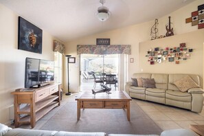 Una sala de estar con televisor, una mesa de centro de madera, un sofá color crema y una zona de comedor con sillas y una mesa.