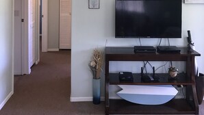 A living room with a flat-screen TV on a wooden stand, a decorative wall piece, and a vase with dried plants.