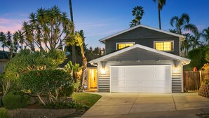 A modern two-story house with a garage, surrounded by lush tropical plants and palm trees.