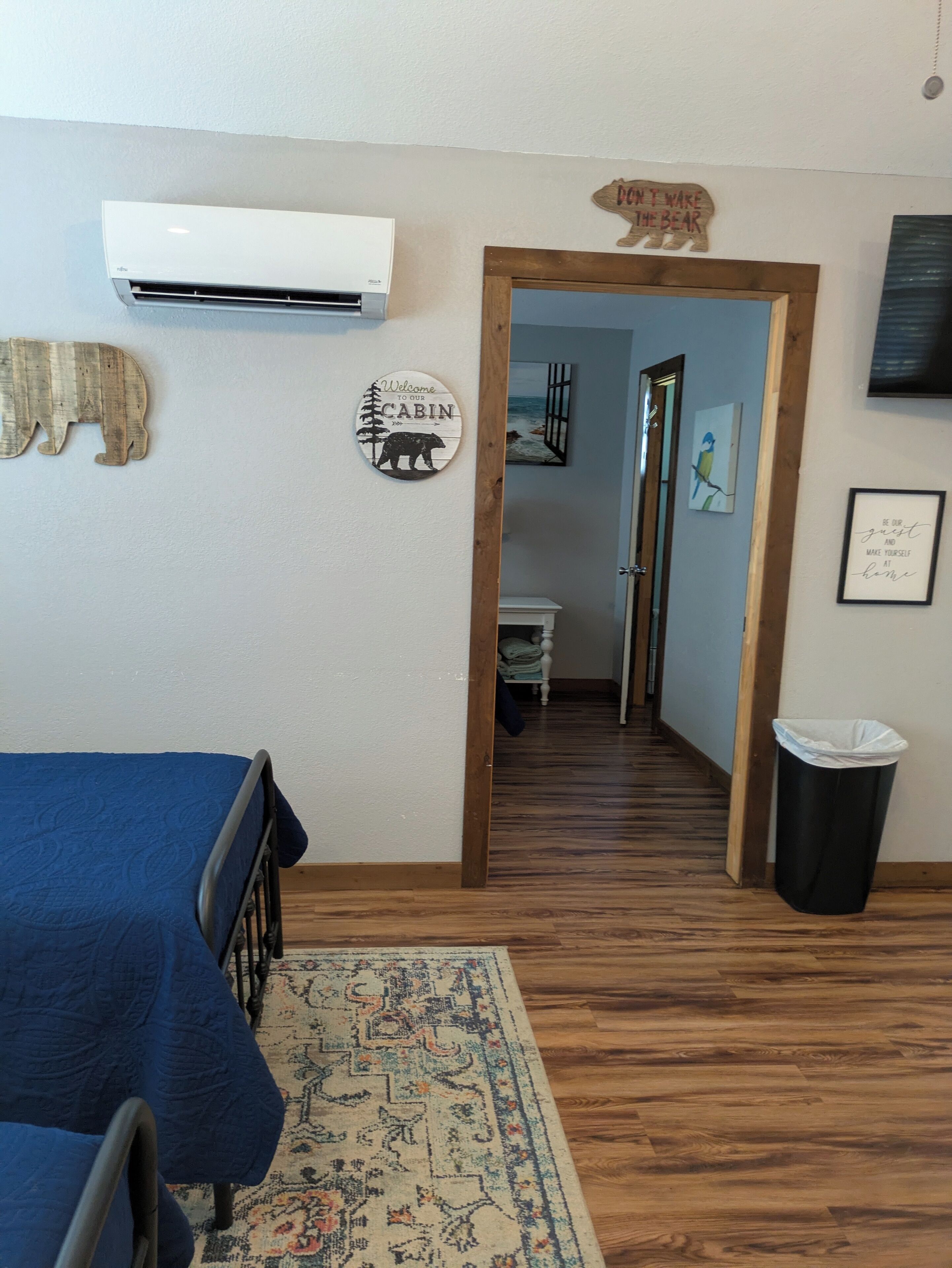 A room with a bed, a wooden floor, a rug, a wall-mounted air conditioner, a wooden door, and a trash bin.