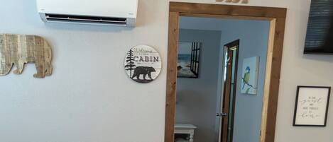 A room with a bed, a wooden floor, a rug, a wall-mounted air conditioner, a wooden door, and a trash bin.