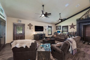 A living room with a brown sofa, a coffee table, and a TV. There are two ceiling fans, a wooden door, and a star decoration on the wall. - Beautiful custom barndominium perfect for Aggie game day, events and getaways! (Caldwell)
