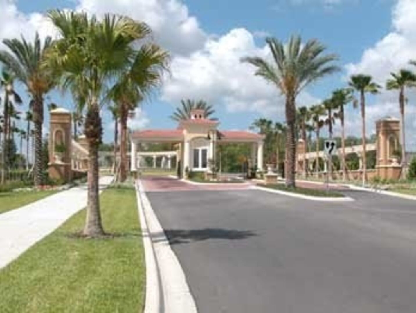 A gated entrance with palm trees, a covered walkway, and a building with a red roof.