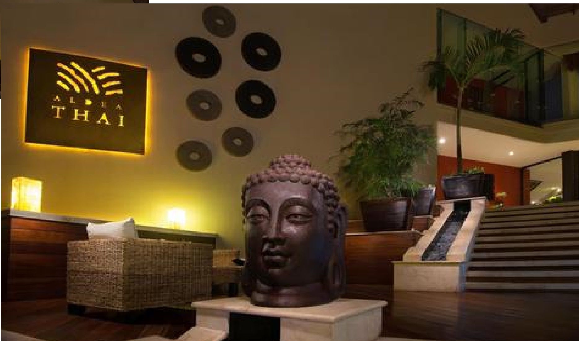 Un espace d’accueil agrémenté d’une sculpture représentant une tête de Bouddha, d’une enseigne murale portant l’inscription « ALPHA THAI » et d’un escalier menant à un niveau supérieur.