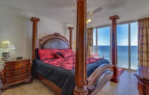 A four-poster bed with a headboard, a nightstand with a lamp, and a view of the ocean through large windows.