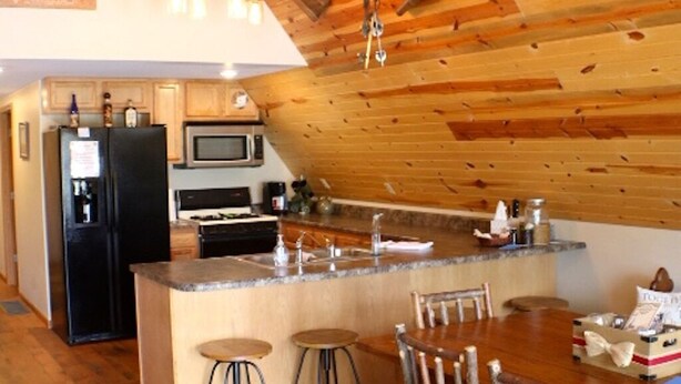 A cozy kitchen with wooden beams, a central island, bar stools, and a dining table. - Cozy A-Frame house in heart of the midwest!!! (Quincy)