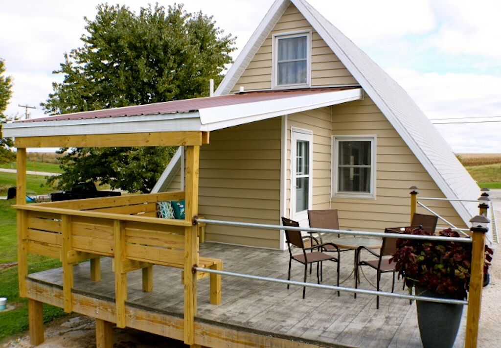 A wooden deck with a covered seating area, a small table, and chairs in front of a house with a gabled roof and a window.