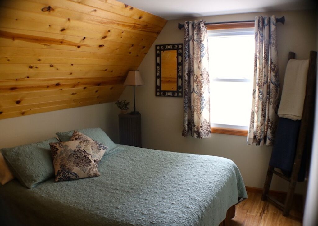 A bedroom with a sloped ceiling, a bed with patterned pillows, a nightstand with a lamp, and a window with curtains.
