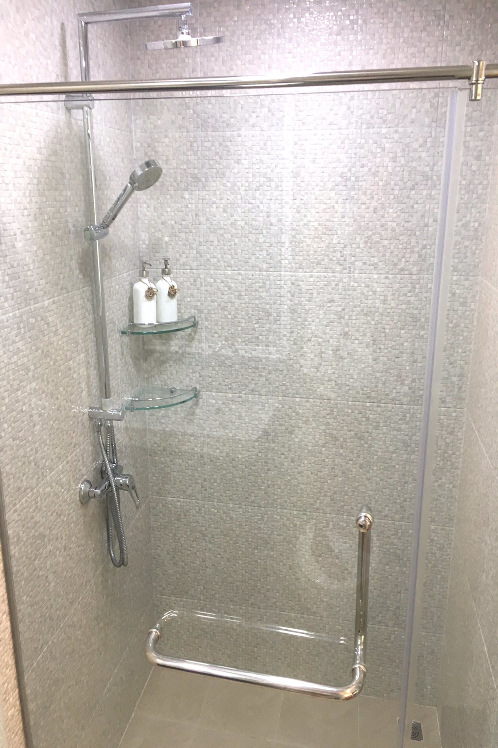 Une cabine de douche fermée par des parois en verre, dotée d’un pommeau de douche à main, d’un flexible de douche et d’une étagère en verre pour les articles de toilette.