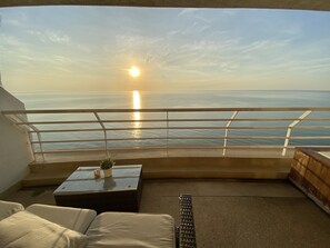 A balcony with a view of the sea and the sun setting. There is a white railing, a small table with a plant, and a sofa. - Sandy Beach Condo 17D (Cha-am)