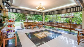 A spacious indoor area with a tiled floor, wooden benches, a chandelier, and a view of greenery outside.
