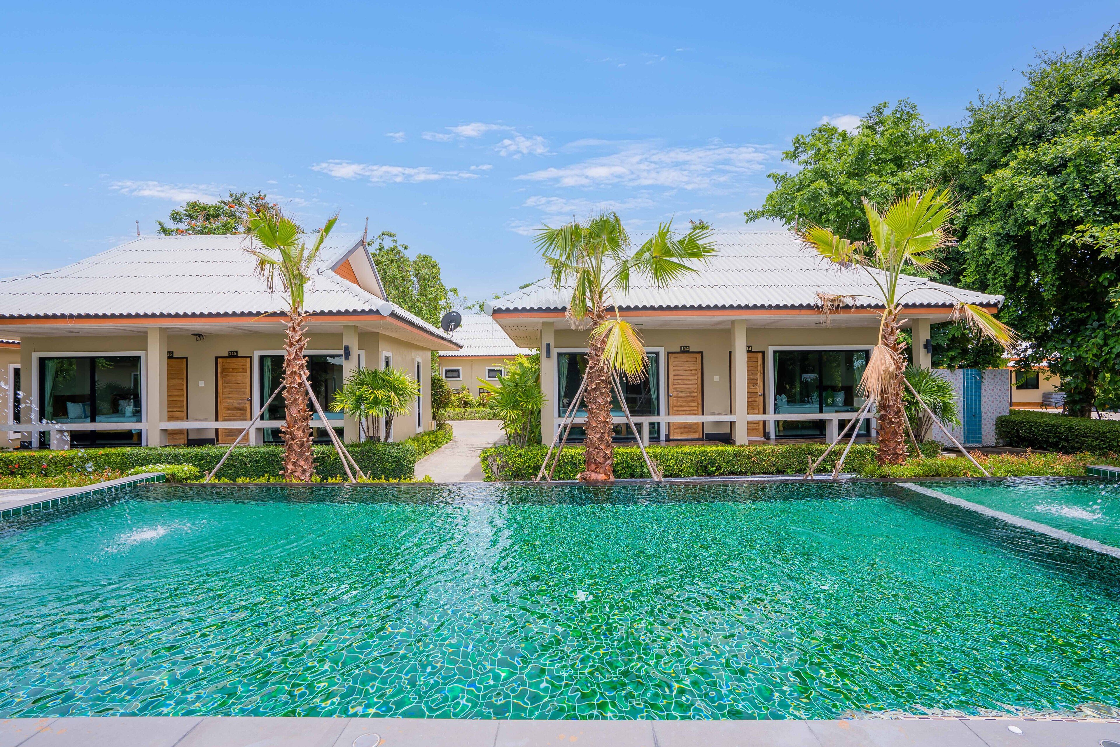 A swimming pool with clear water, surrounded by two buildings with tiled roofs and palm trees.