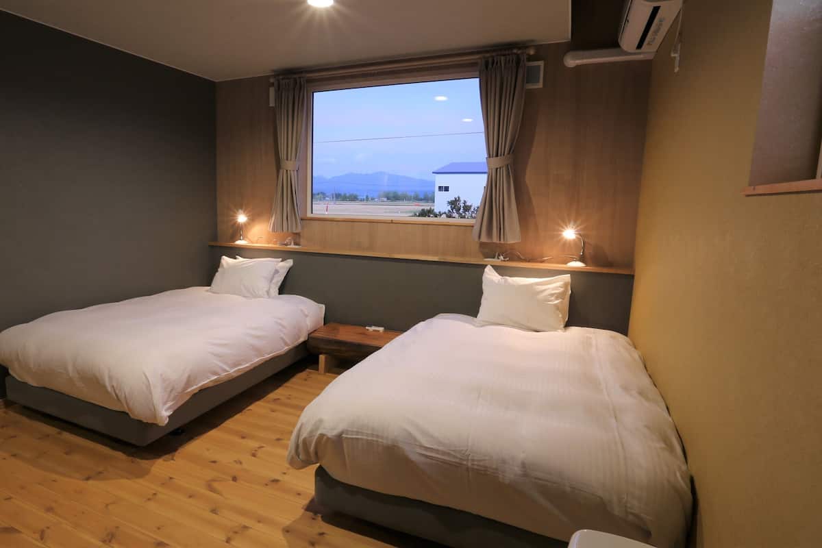 Una camera da letto con due letti, pavimento in legno, una finestra con tende e una luce notturna.