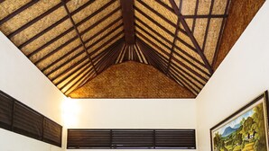 A room with a high, sloped ceiling made of wooden beams and thatch, a dark wooden window shutter, and a framed landscape painting on the wall. - Murni's Villa Kelor (Ubud)