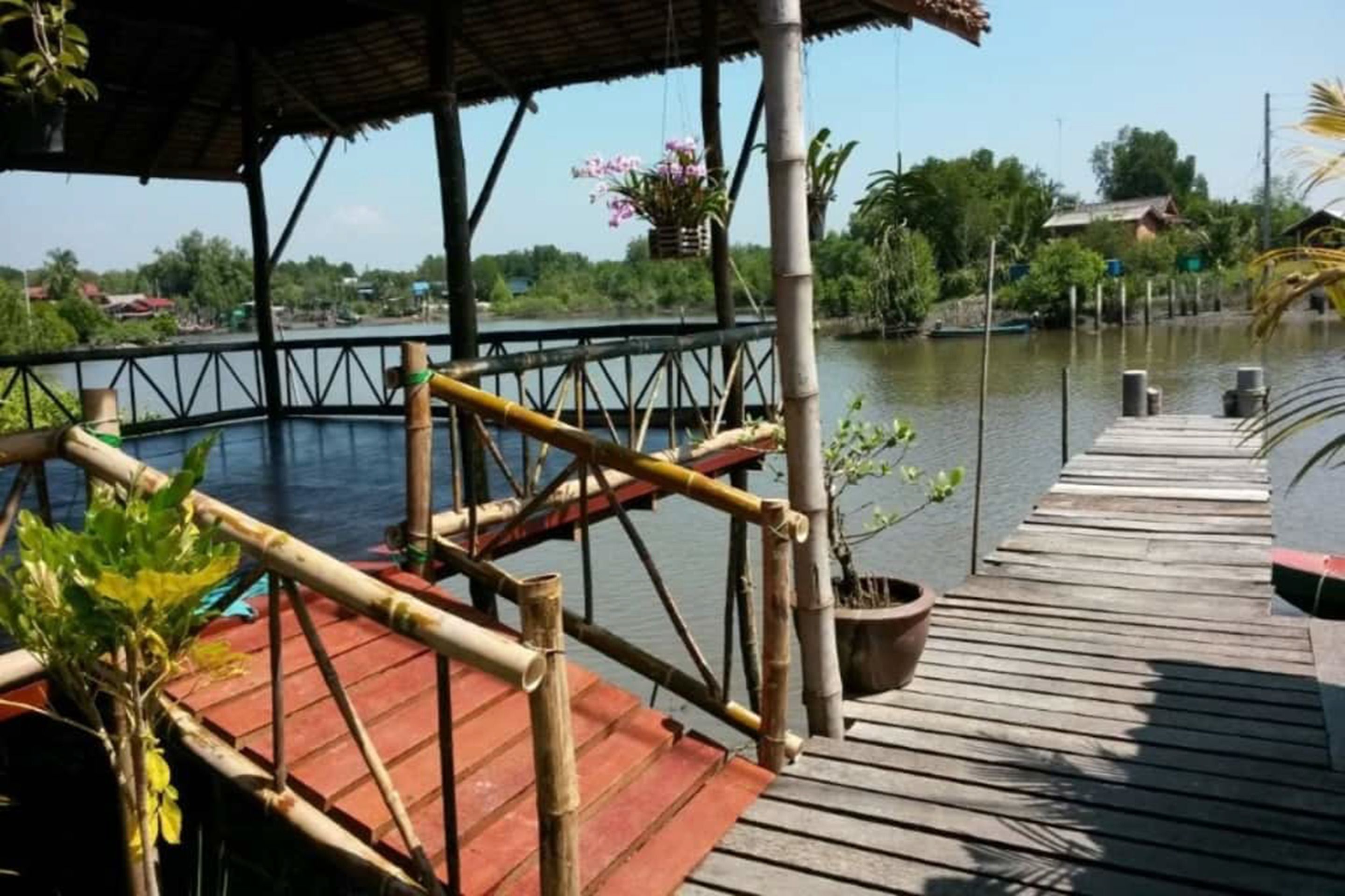 Un muelle de madera que se extiende sobre el agua, con baranda de bambú y un área cubierta decorada con flores colgantes.