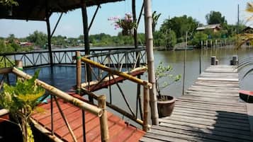 Un muelle de madera que se extiende sobre el agua, con baranda de bambú y un área cubierta decorada con flores colgantes.
