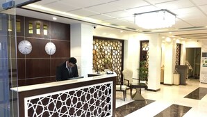 Un espace d’accueil d’hôtel comprenant un comptoir de réception avec un réceptionniste, deux horloges murales, un élément de séparation décoratif et un lustre moderne.