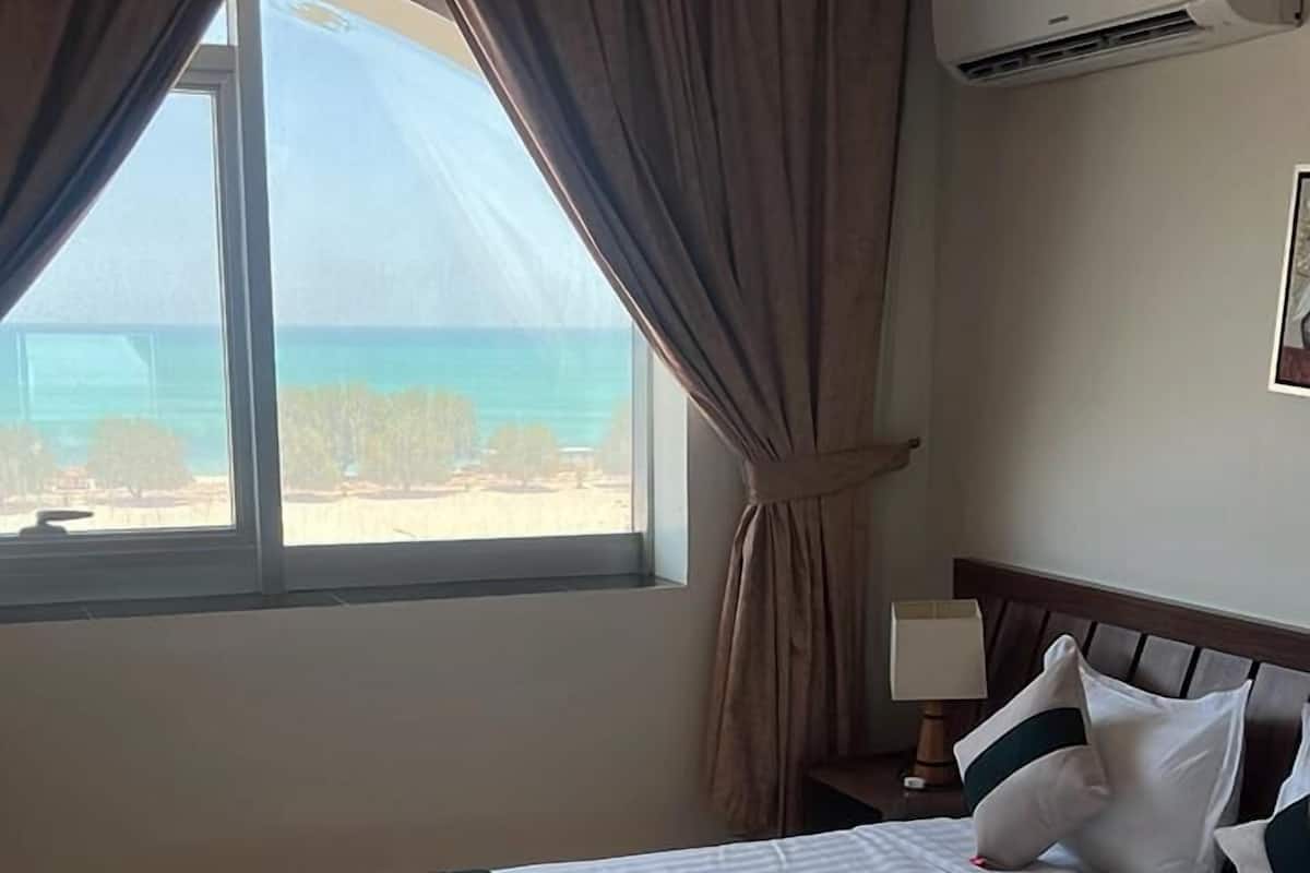 A hotel room with a large bed, a nightstand, a lamp, and a view of the beach.