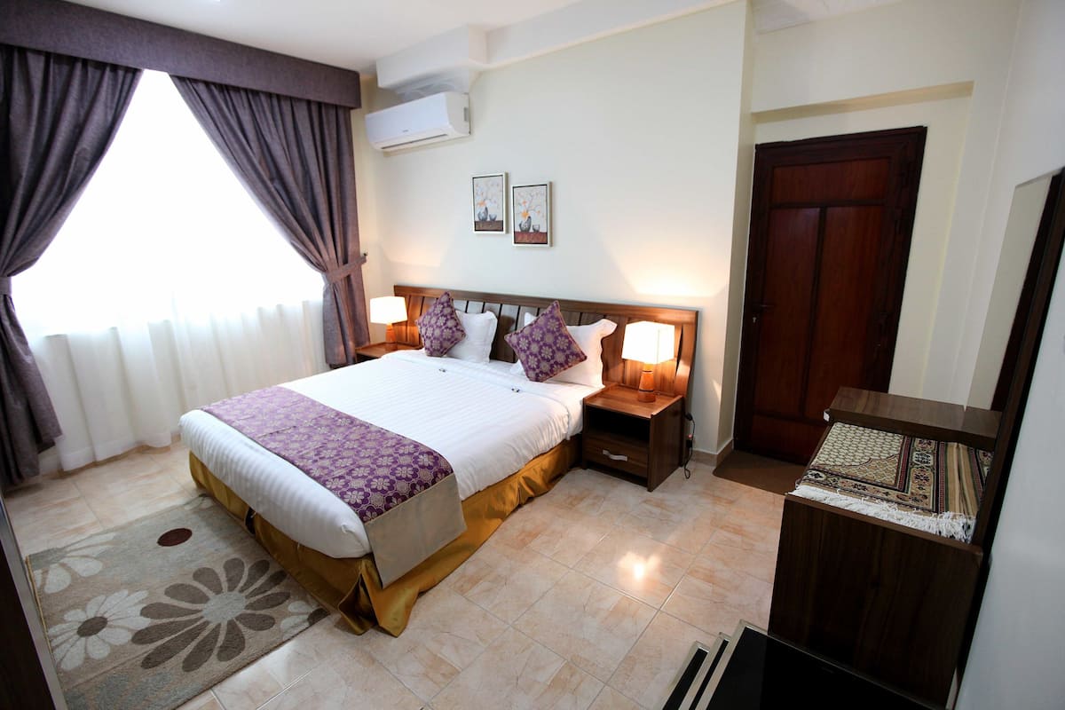 A neatly arranged bedroom with a large bed, bedside lamps, a wooden nightstand, a door, and a wall-mounted air conditioner.