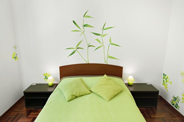 Un lit bien fait, avec une couverture verte et des oreillers, deux tables de chevet avec des lampes, et un mur décoré d’un motif représentant une plante de bambou.