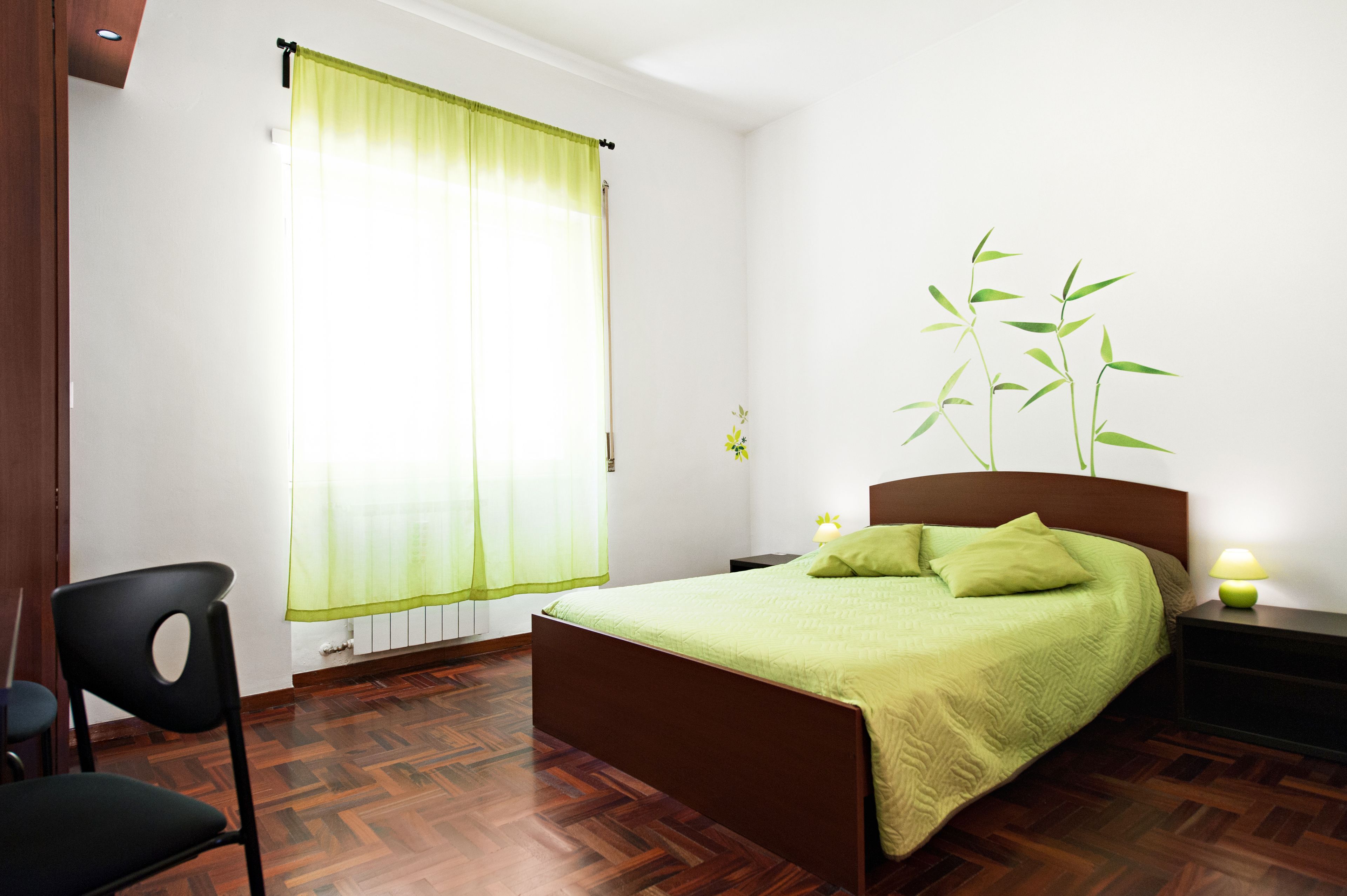 A bedroom with a bed, a chair, a nightstand, a lamp, and a window with a curtain.