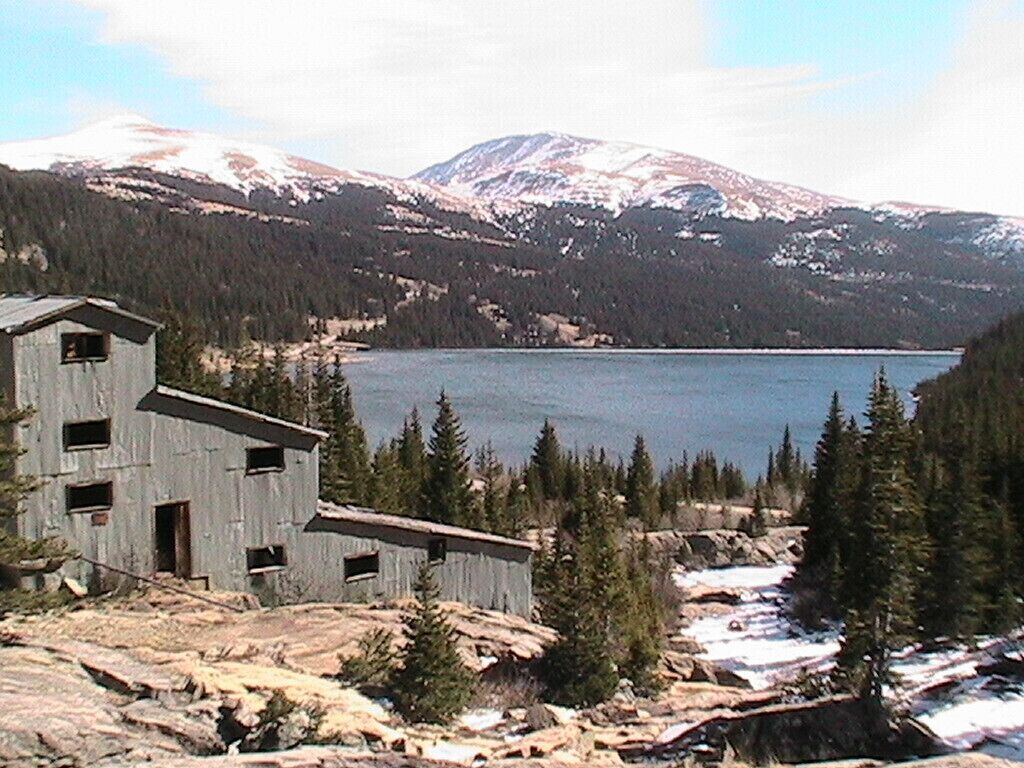 A rustic cabin with a small porch, surrounded by a forest and a lake, with snow-capped mountains in the background.