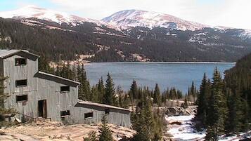 A rustic cabin with a small porch, surrounded by a forest and a lake, with snow-capped mountains in the background.