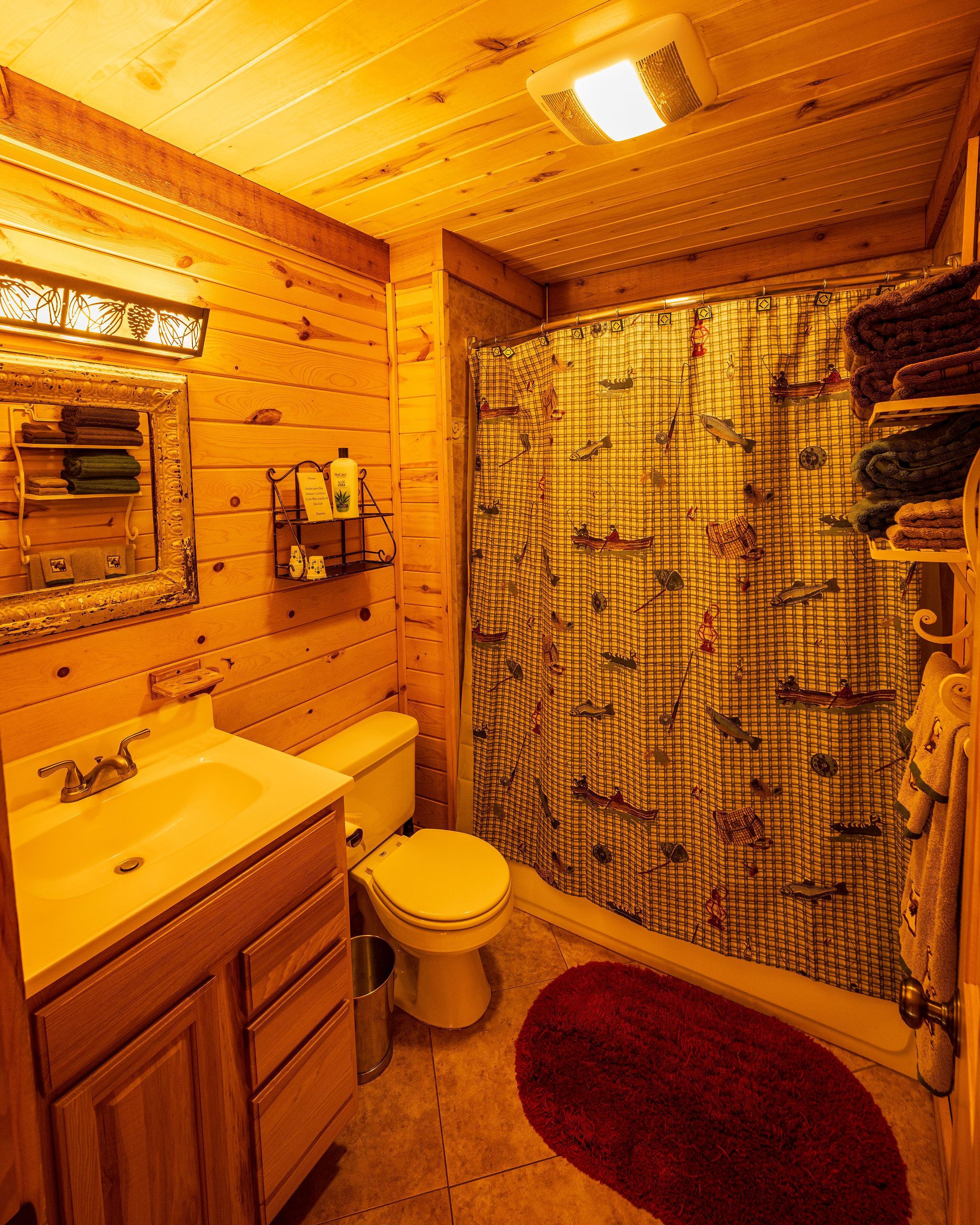 Une salle de bain rustique, aménagée dans une cabane en bois, avec un lavabo, des toilettes et un rideau de douche orné d’un motif de poissons.