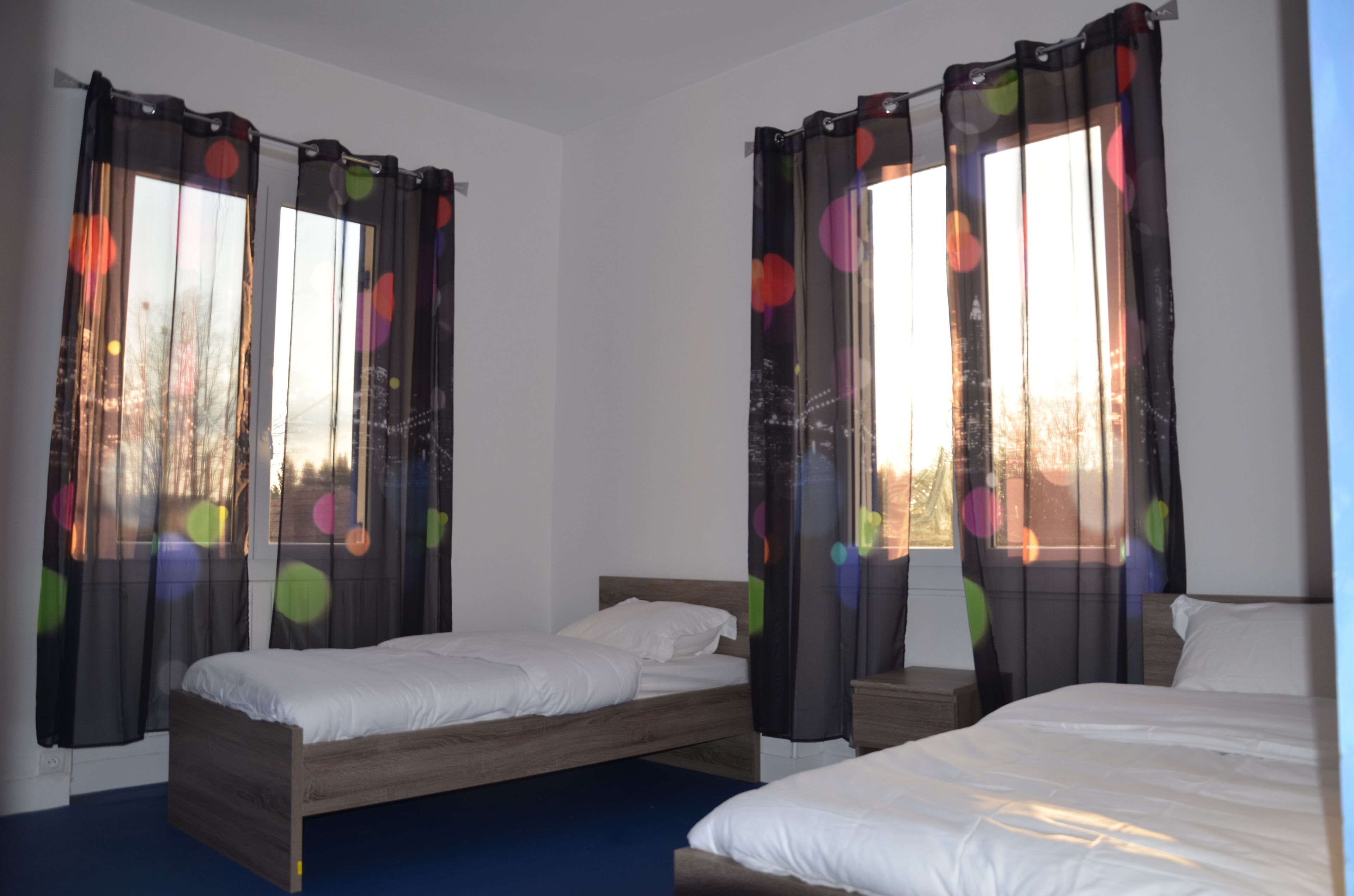 A room with two single beds, a wooden headboard, and sheer curtains with colorful polka dots.