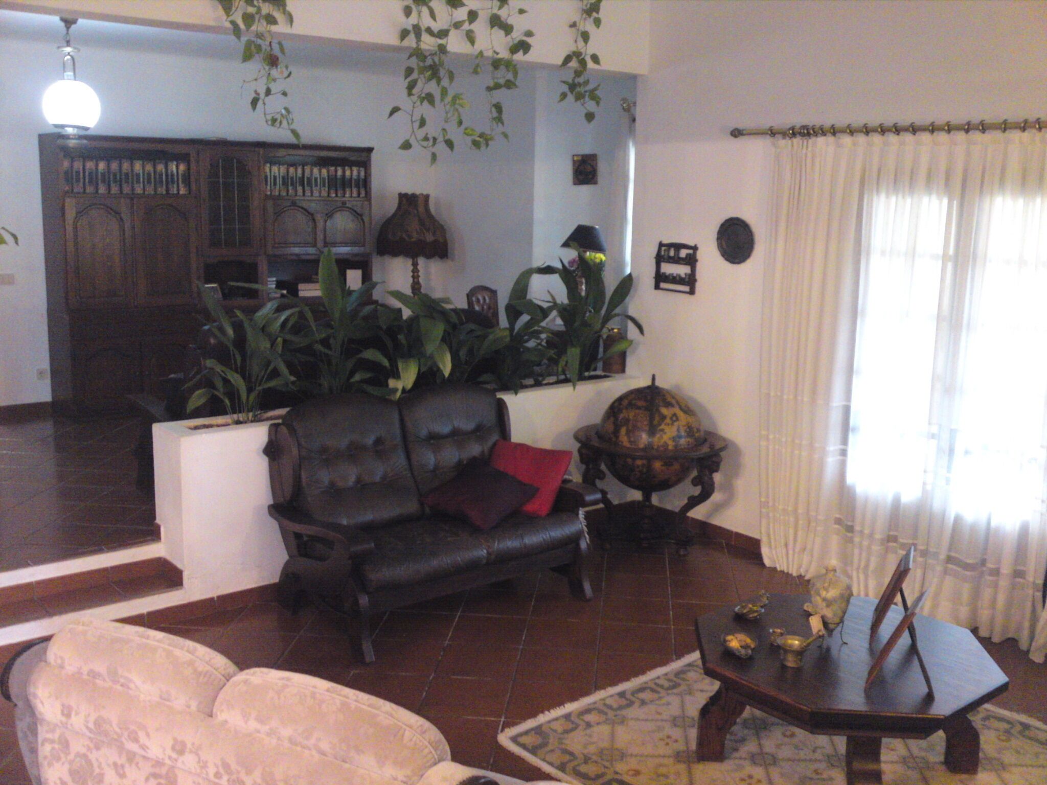 A living room with a sofa, a coffee table, and a lamp. There are plants and a cabinet in the background.