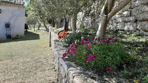 Un muro de piedra con un jardín lleno de flores, un árbol y un edificio con techo de tejas.