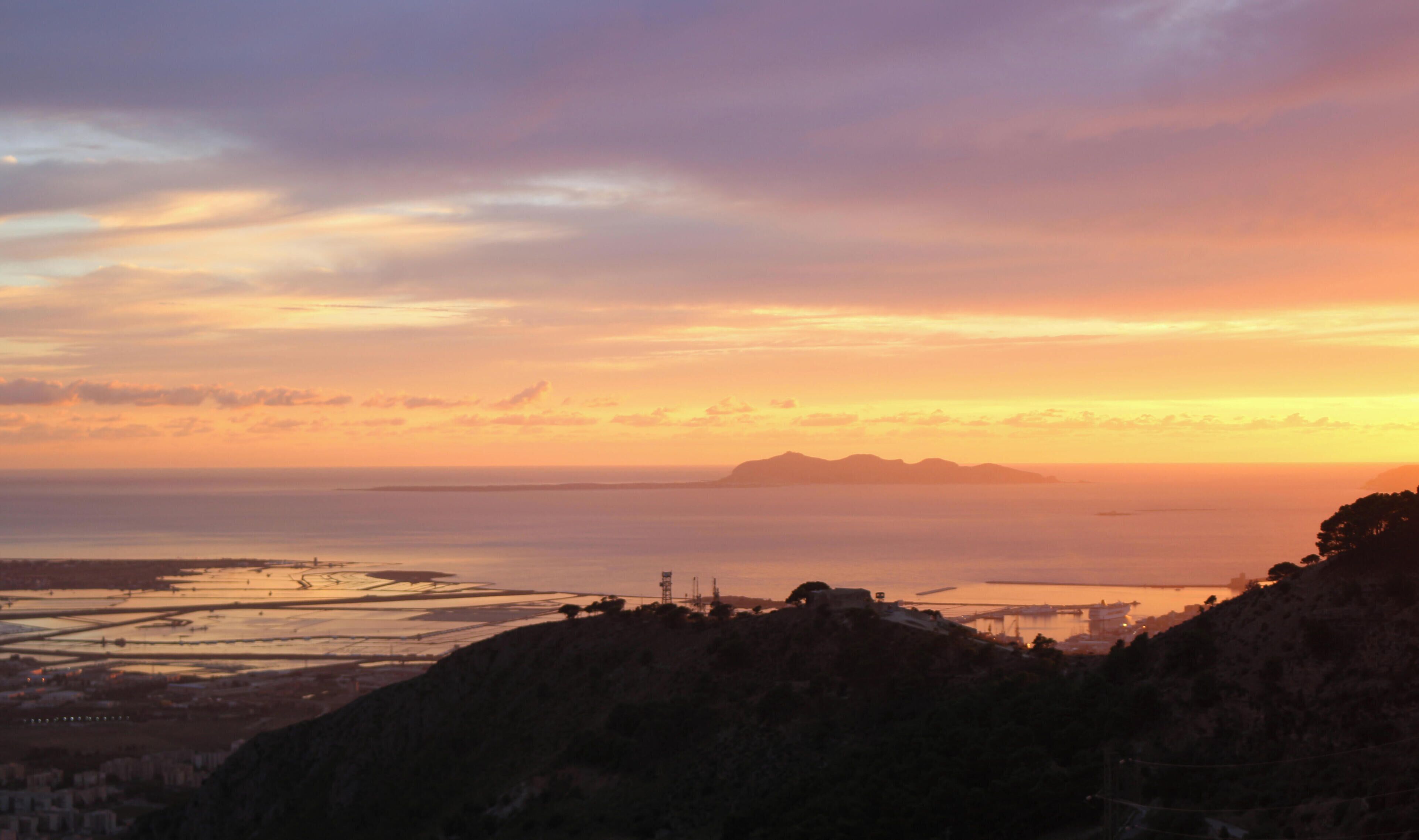 A coastal landscape with a sunset, featuring a prominent hill in the foreground and a distant view of a bridge and water.
