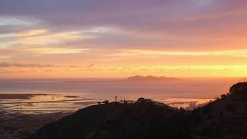 A coastal landscape with a sunset, featuring a prominent hill in the foreground and a distant view of a bridge and water.