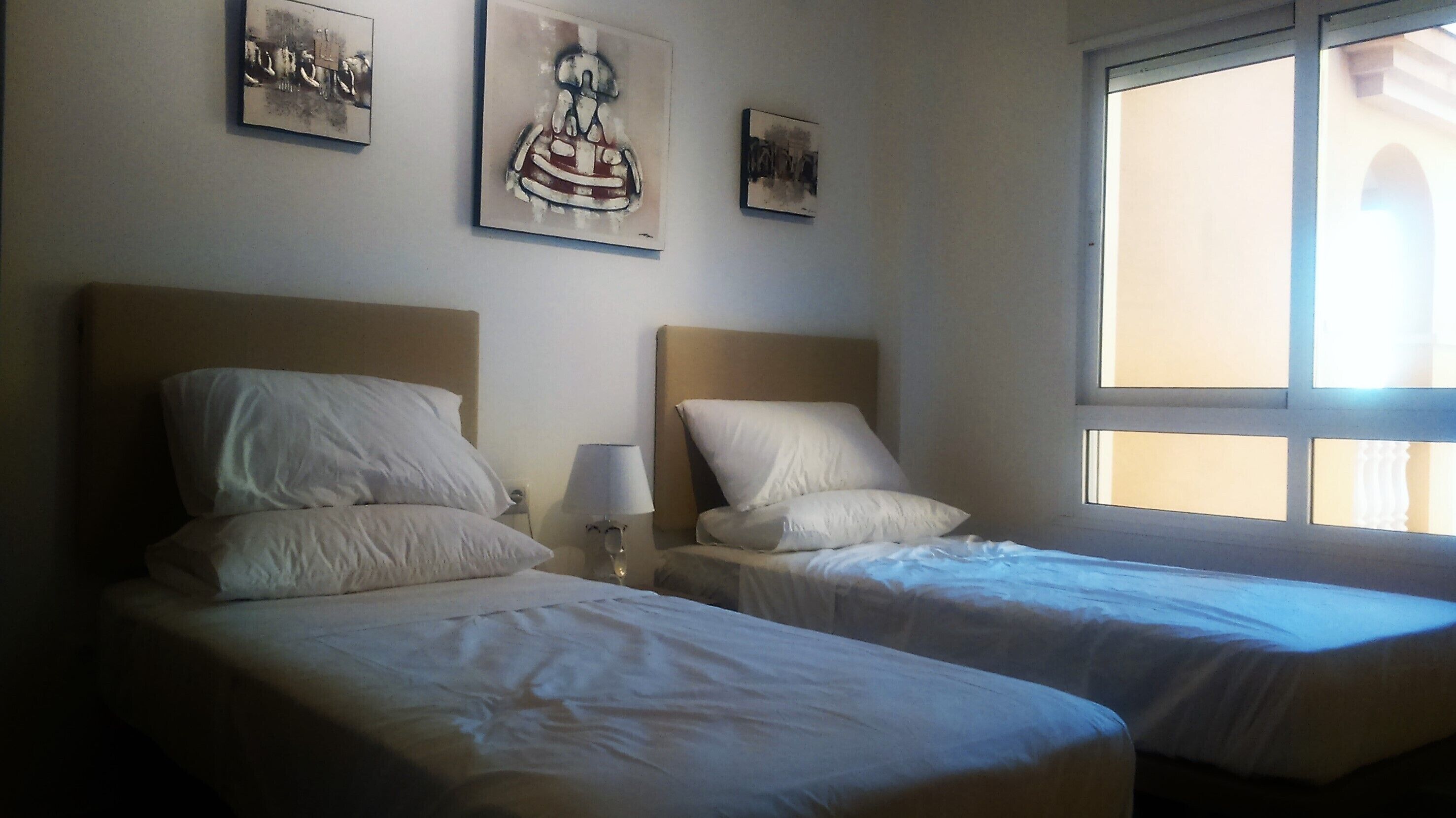 Habitación de hotel con dos camas, cada una con cabecero y almohadas, una mesita de noche con lámpara y una obra de arte en la pared.