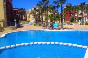 A swimming pool with white stepping stones, surrounded by palm trees and a building with balconies.