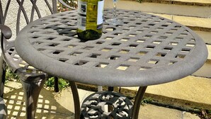 Een ronde metalen tafel met een fles witte wijn en een glas, buiten geplaatst met een huis op de achtergrond.