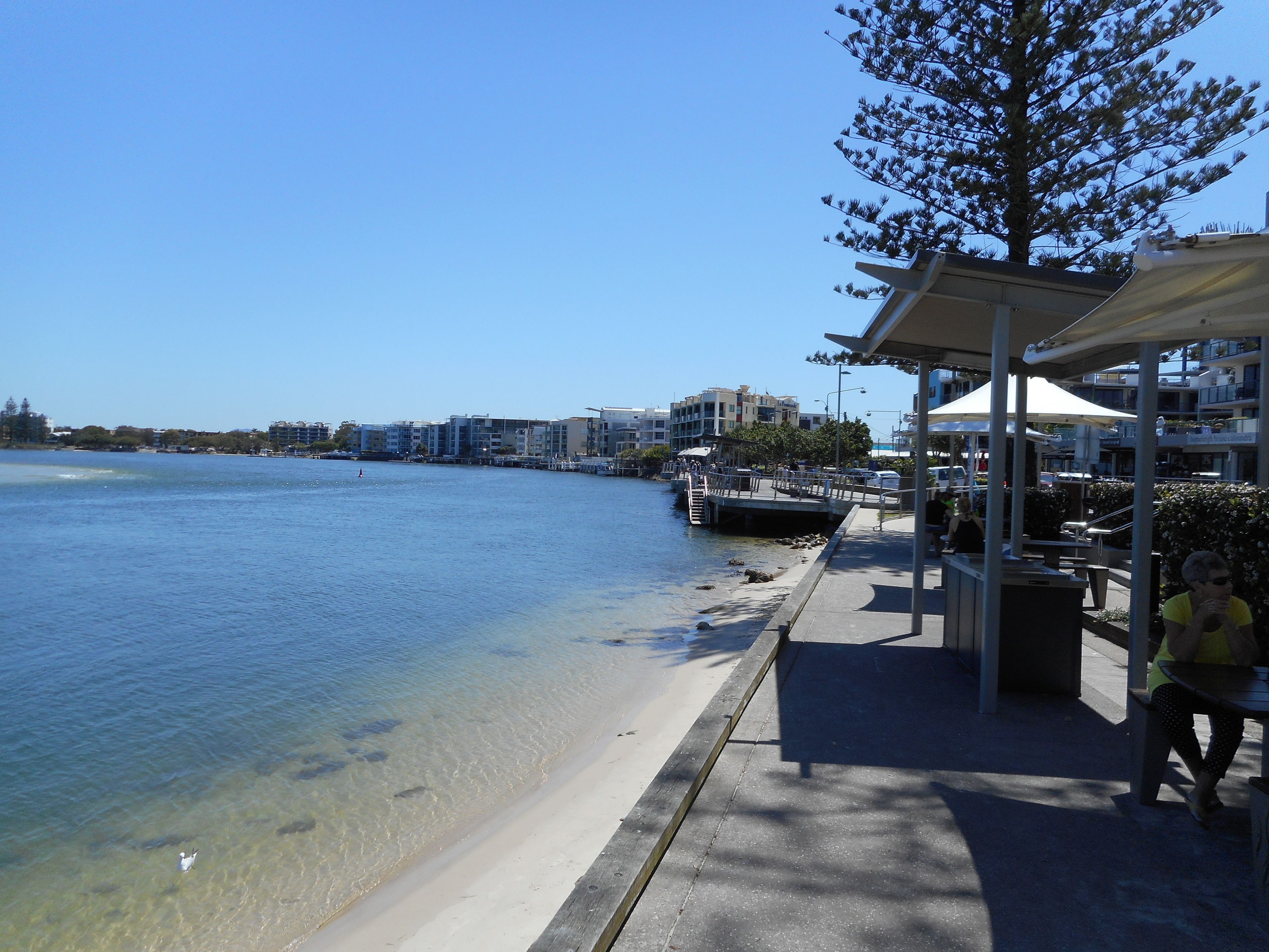 A waterfront promenade with a clear view of the sea, a covered seating area, and buildings in the background.