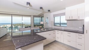A modern kitchen with a large island, white cabinetry, and a view of the ocean. - The Waterford Prestige Apartments (Caloundra)