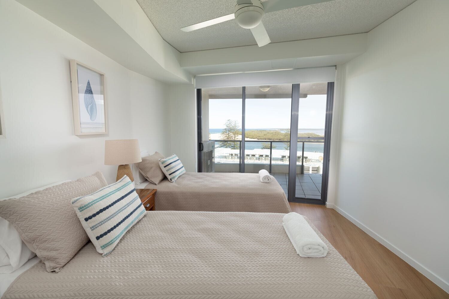 A modern hotel room with two beds, a ceiling fan, a large window with a view of the sea, and a balcony with outdoor furniture.