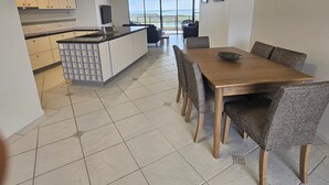 A modern kitchen with white cabinets, a central island, and a dining area with a wooden table and wicker chairs. - The Waterford Prestige Apartments (Caloundra)