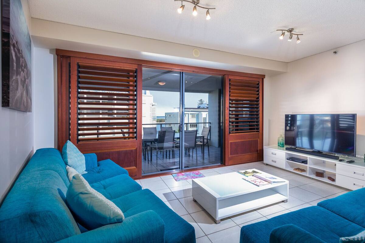 Una sala de estar moderna con un sofá azul, un televisor de pantalla plana y un ventanal corredero que da a un balcón exterior.