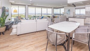 A spacious living area with a white sofa, wicker chairs, and a dining table. Large windows offer a view of the city and greenery. - The Waterford Prestige Apartments (Caloundra)