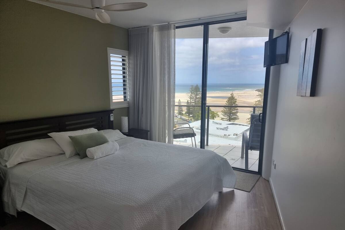 Habitación de hotel con cama, ventilador de techo, televisor montado en la pared y balcón con vista a la playa.
