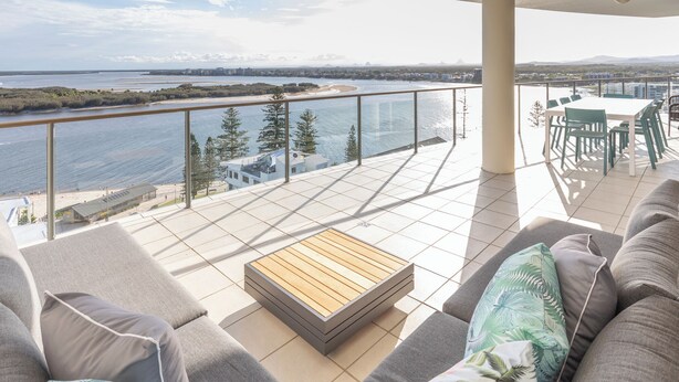 A balcony with a view of the sea, a wooden table, and grey sofas with tropical pillows. - The Waterford Prestige Apartments (Caloundra)