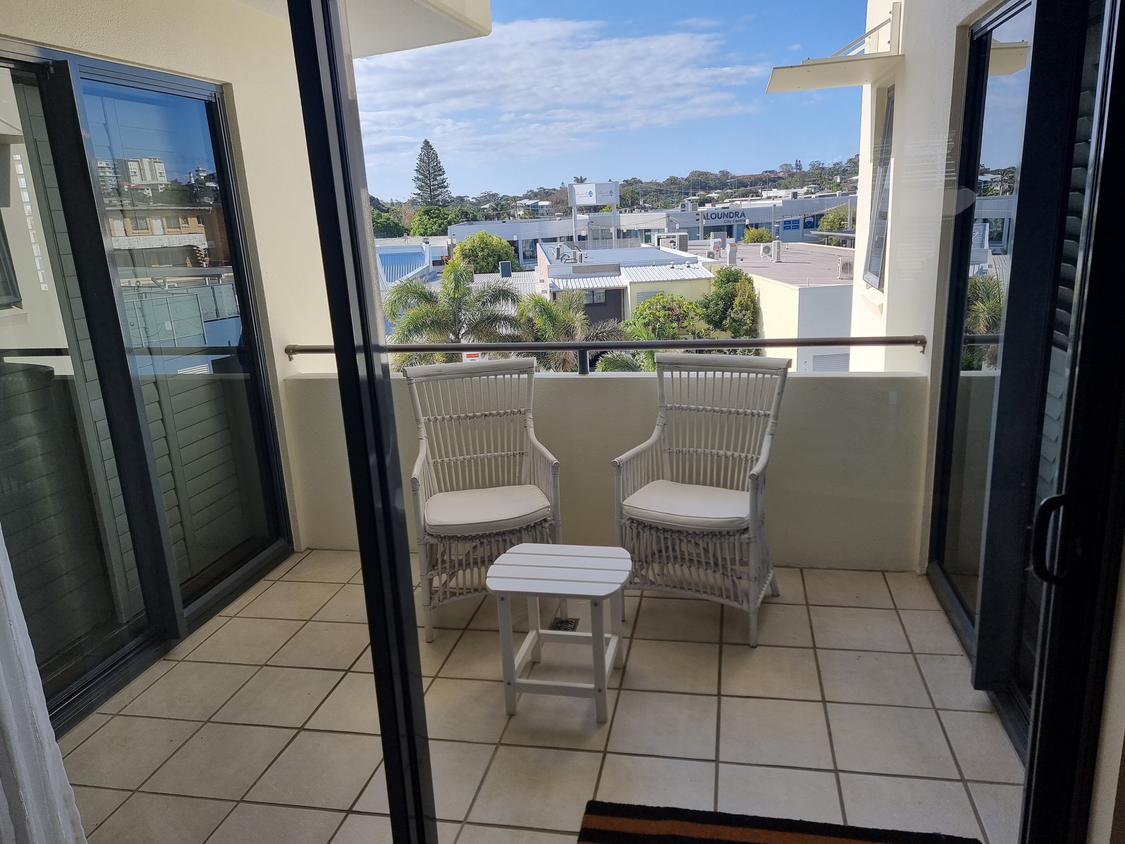 Un balcon avec deux fauteuils en osier et une petite table, d’où l’on peut admirer un panorama urbain avec ses bâtiments et sa verdure.