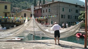 A man standing in front of a netted area by a body of water, with buildings and a mountainous backdrop. - Great Price... Romantic Apartment Near Beaches and Nightlife (Malcesine)