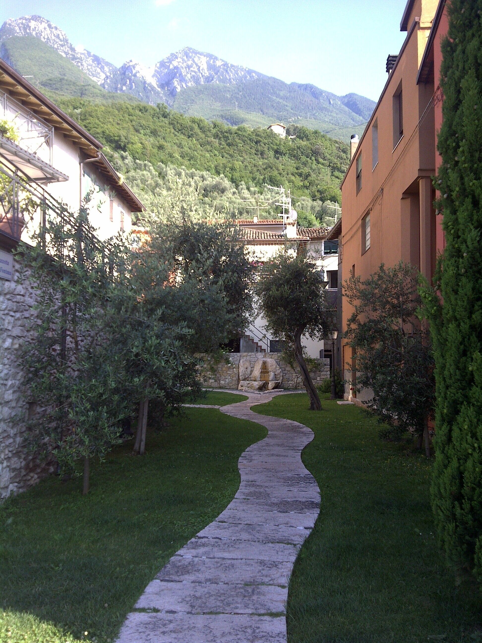 A stone pathway leading through a courtyard with buildings on either side, surrounded by greenery and mountains in the background.